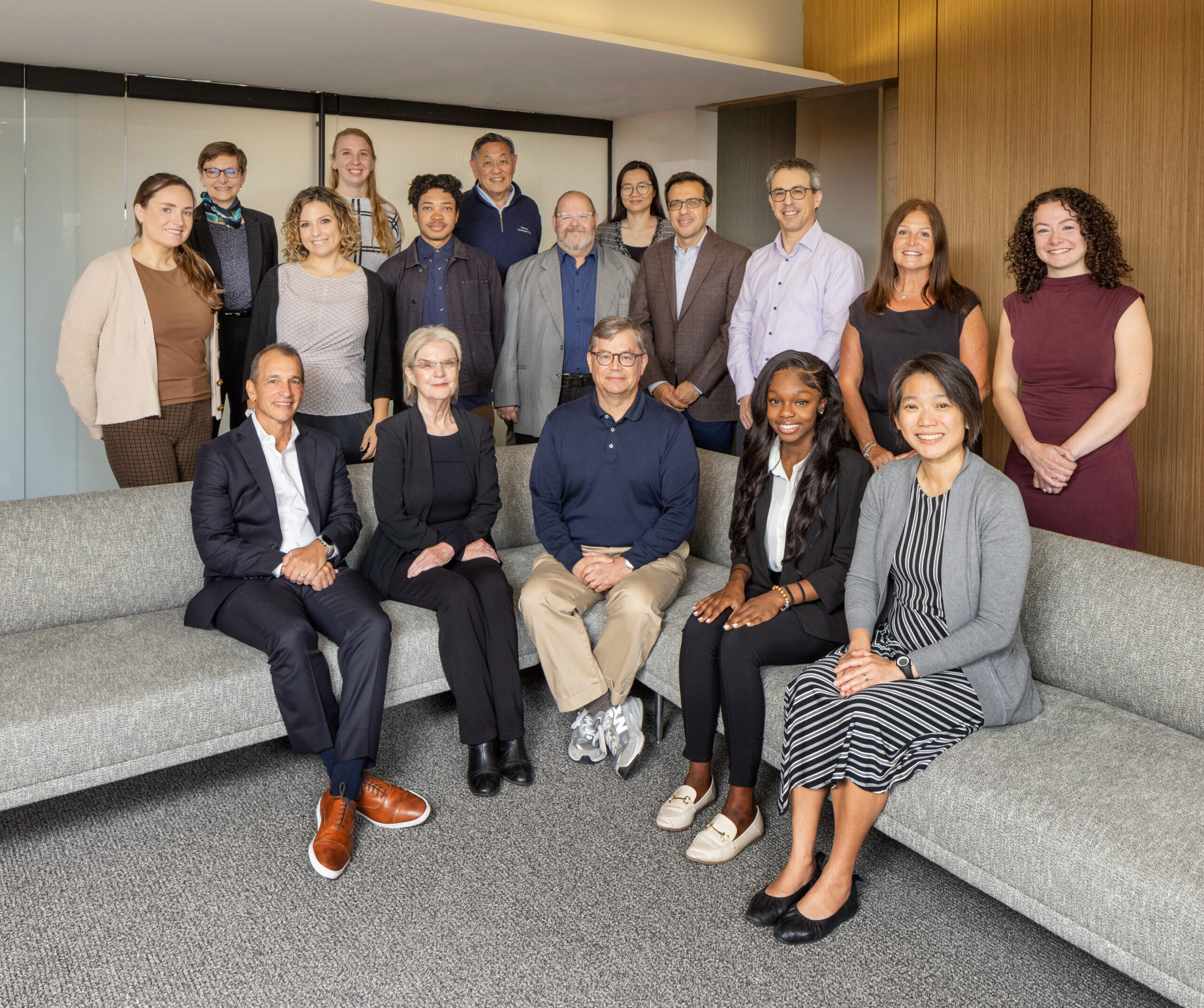 A group of people posing for a professional group photo in an office setting. They are dressed in business and business-casual attire, standing and sitting on a couch.